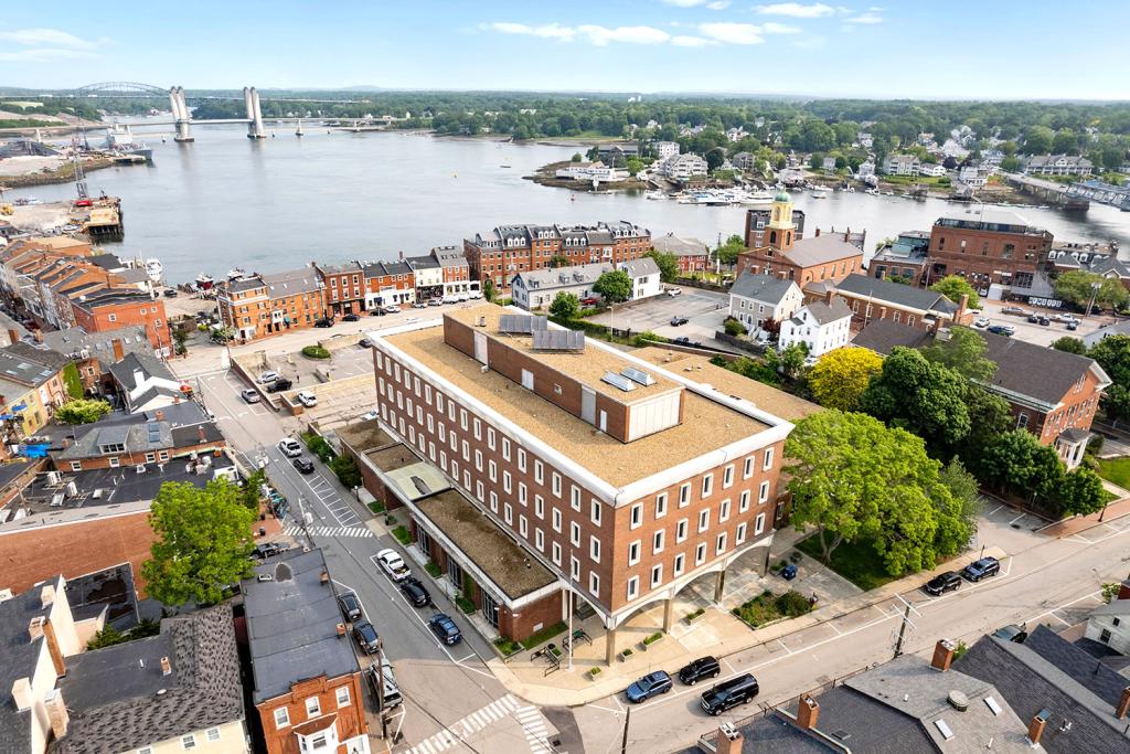 Aerial image of a four-story red brick building on a corner overlooking a parking lot, a row of brick buildings, streets, a harbor, and a bridge. A portico faces a sidewalk.