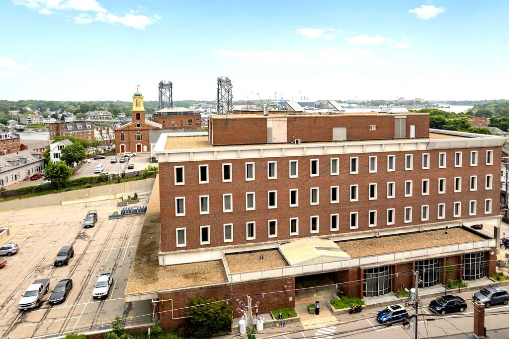 Image of a four-story red brick building with a large one-story annex and a parking lot. A bridge and a church are in the background.