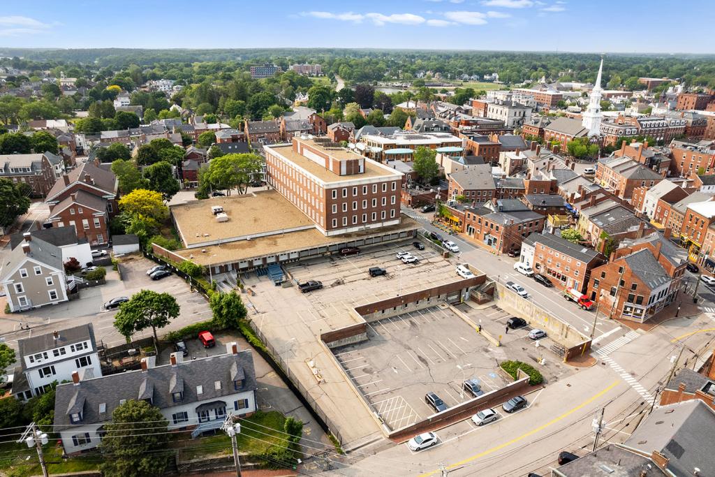 Aerial image of a four-story red brick building with a large one-story annex overlooking a two-level parking lot with loading docks and garage. The building sits in the middle of a downtown area with a white church and red brick buildings.