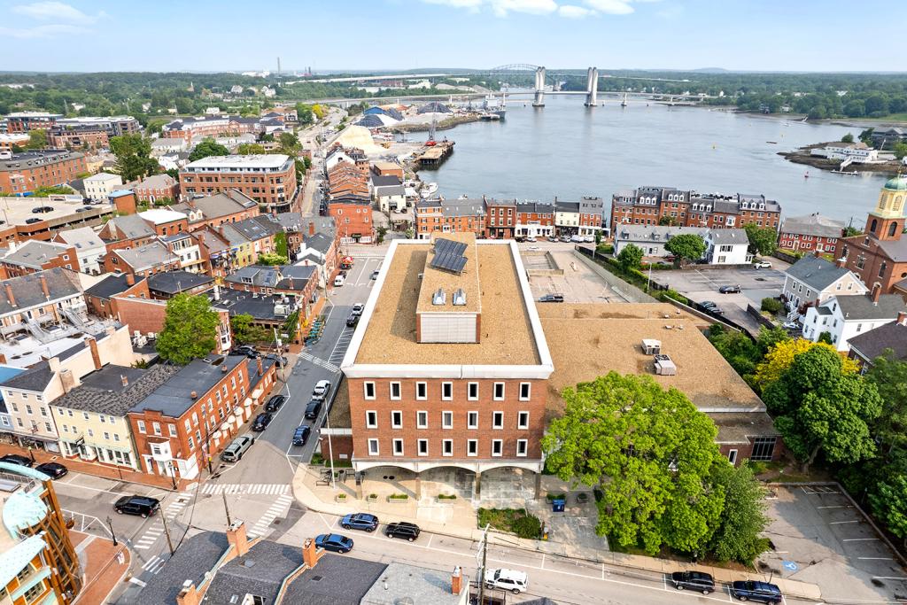 Aerial image of a four-story red brick building with a large one-story annex overlooking a two-level parking lot with loading docks and garage. The building overlooks a row of brick buildings, a harbor,and a bridge. The main building has a portico.