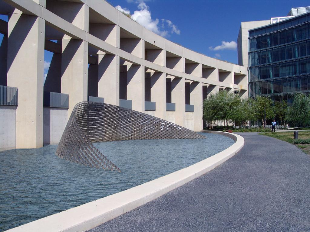 A 100-foot long, wind-animated curved stainless steel sculpture, immersed a reflecting pool between a curved Brutalist architectural wall and a rocky path, with a blue sky