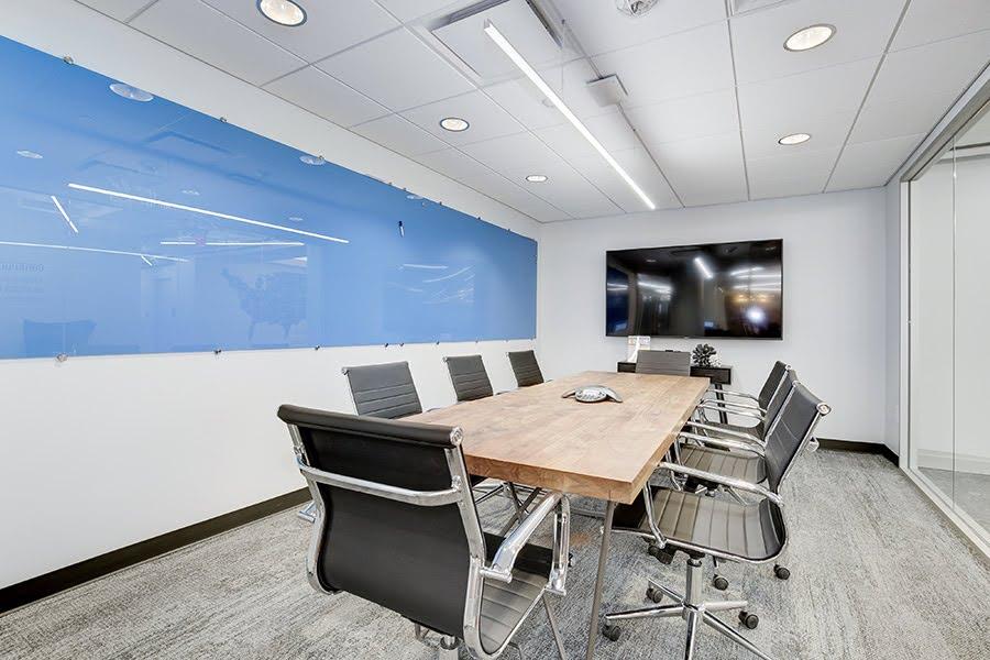 A conference room with eight office chairs around a wood laminate conference table with a glass marker board on one wall and a large flat screen on another wall.
