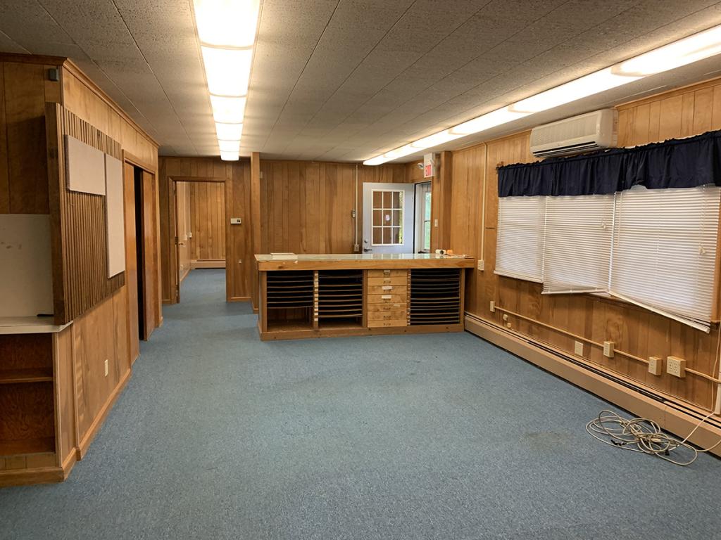 Empty room interior with natural wood paneling, acoustic tile ceiling, blue carpeting, and view into other empty room beyond