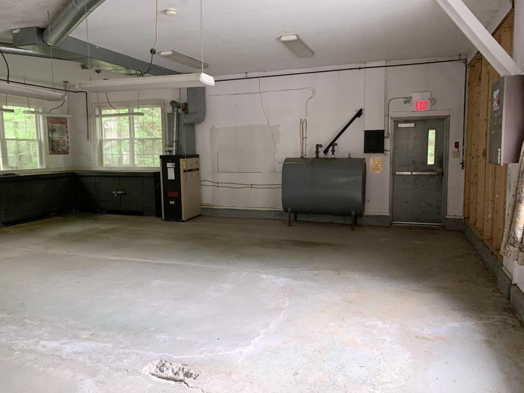 Large, garage-like room interior with white walls, built-in tables along some walls, cement floor, windows, and a gray tank next to an exit door