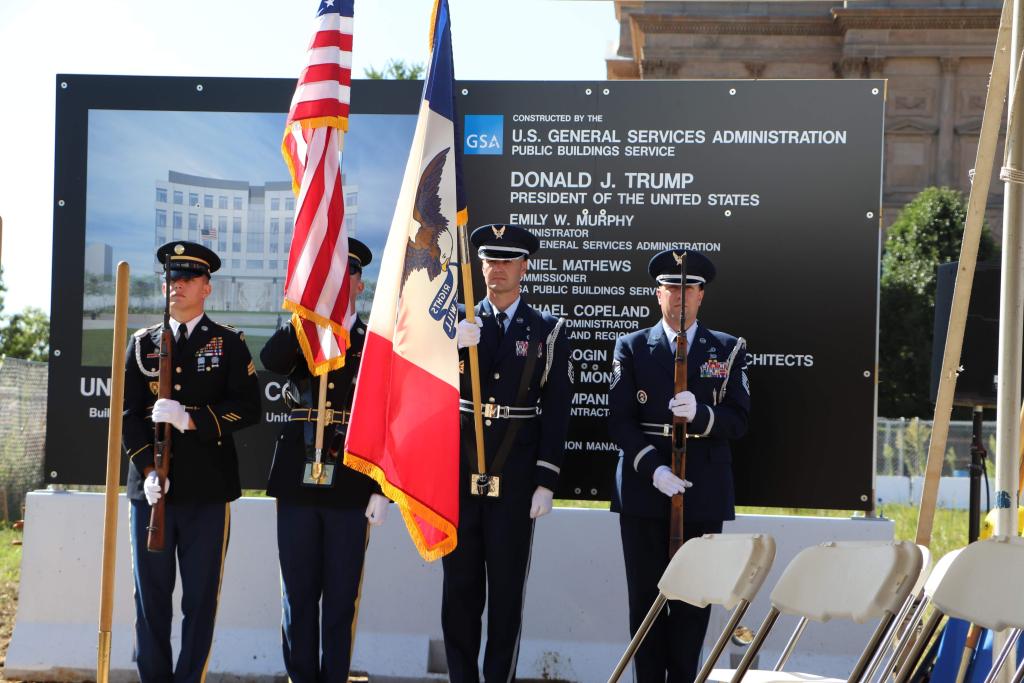Four men stand in military uniforms holding a U.S. flag erect and an Iowa state flag dropped to a 45 degree angle in front of a sign with information about the courthouse construction project