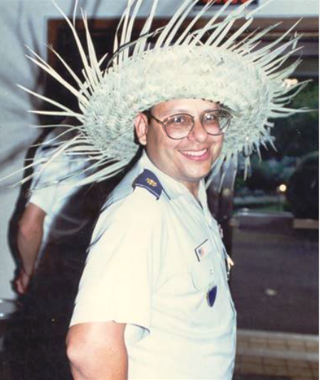 A medium-skinned man wearing a straw sombrero and white collared shirt of a military uniform stands with his arms behind him and looking at the camera over his right shoulder.