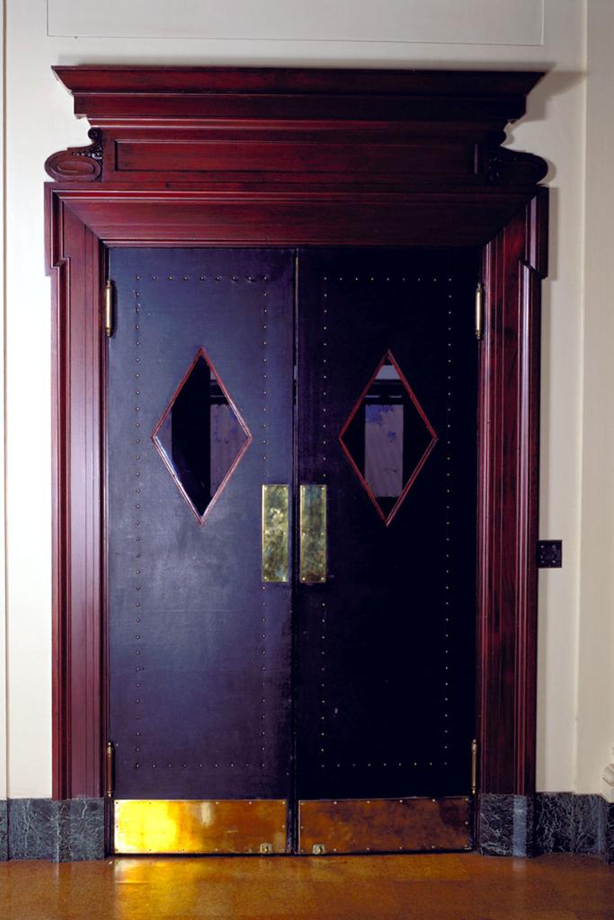 Doorway with two double black swinging doors, each with a diamond-shape window, decorative rivets in an outline, and brass push and kick plates, with an ornate red wood frame surrounding it