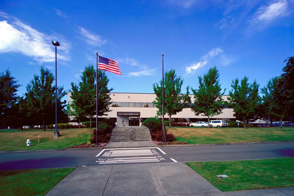 Two-level gray building with blue sky, medium trees at regular intervals, grass, a flag and a road with crosswalk