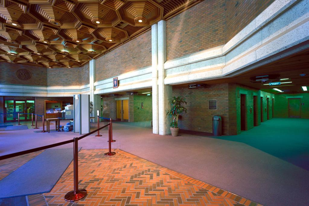 Interior two-story open area of brown brick walls and mauve carpet and red-brick floor, with elevator bank on right, cordoned area on left, and sculptural hexagonal wood ceiling