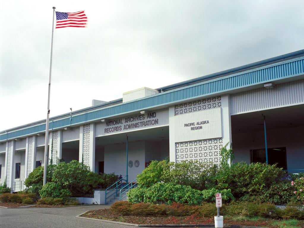 White and blue one-level building entrance with green bushes in front, and National Archives and Records Administration, Pacific Alaska Region signage, and U.S. flag on a pole