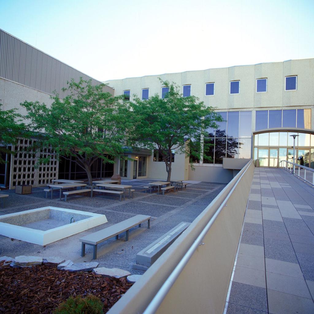 Exterior partial view of multi-section building with glass entrance, and two trees, an empty fountain, and benches on a patio