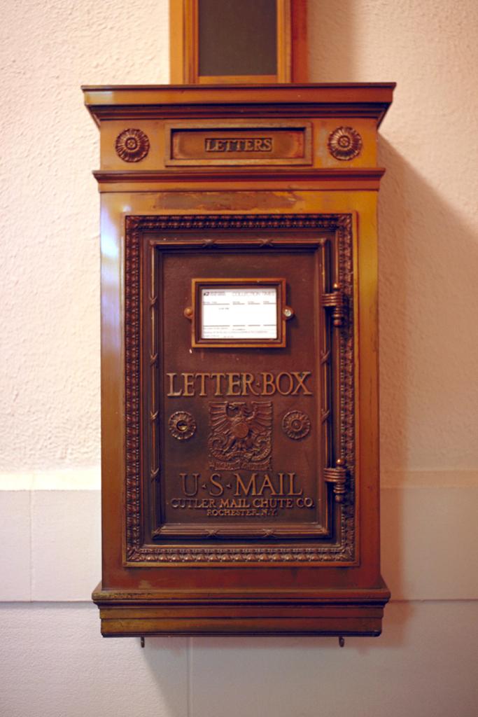 Brown, rectangular, ornate old-fashioned U.S. Mail letter box attached to a tan wall