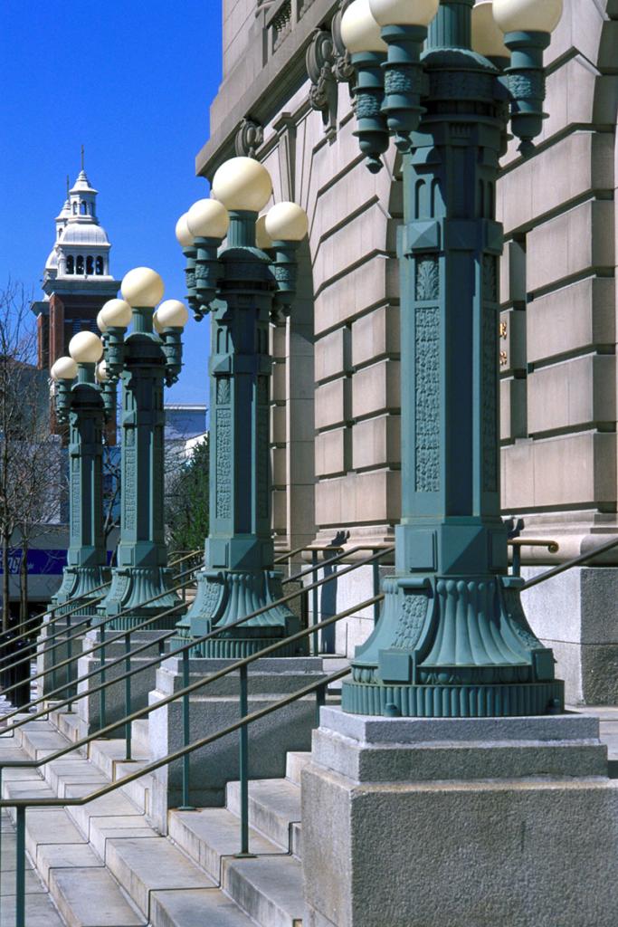 Angled view of front of a cement building with four ornate green lampposts in-between stairs and railings, with a red tower in the distance and blue sky