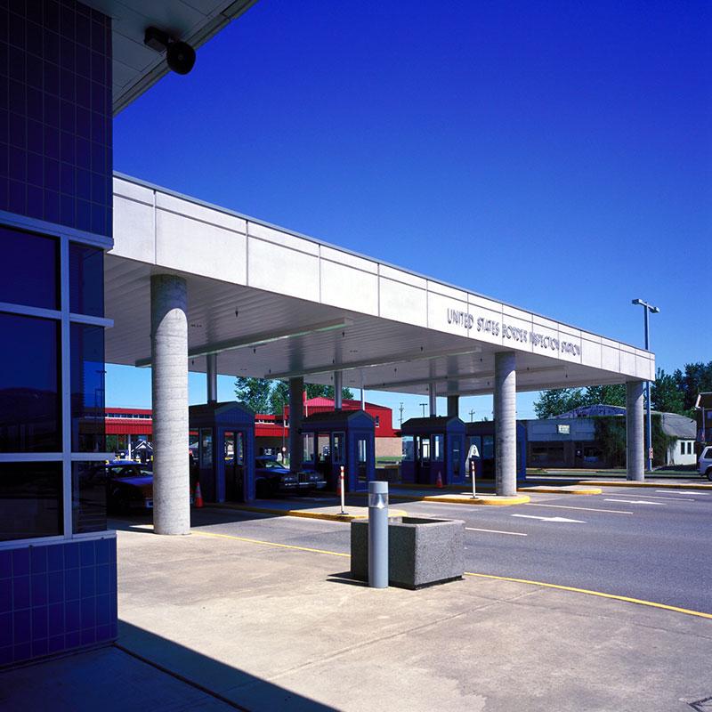 Angled view of an inspection station, with a cover supported by columns, with the words United States Inspection Station on it, four pavement lanes, each with a blue booth, with blue sky in the background