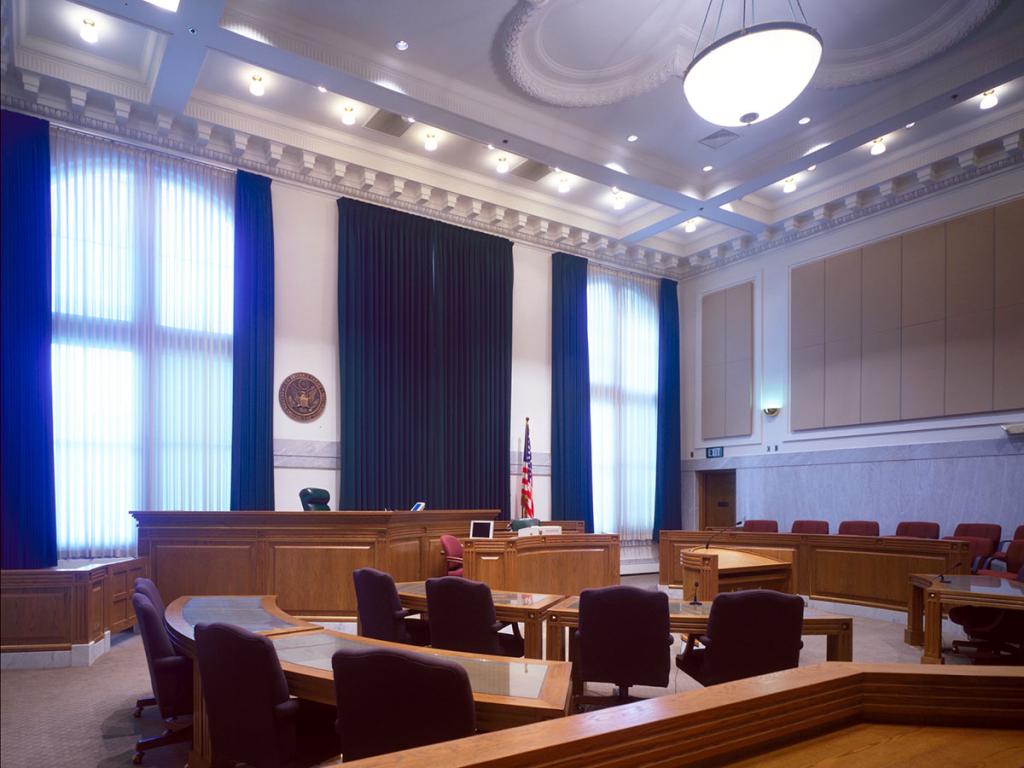 Courtroom interior with blond wood desks, benches and tables, a high ceiling with a round hanging lamp, and a wall of windows with blue and white curtains