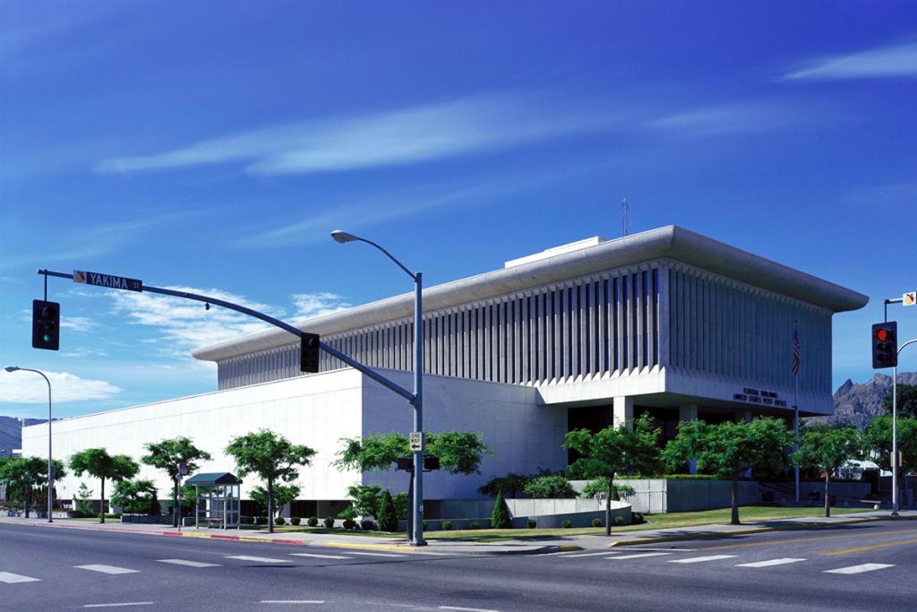 Angled view of a white, multi-level concrete building with surrounding trees, bushes, roads, traffic signals and a blue sky