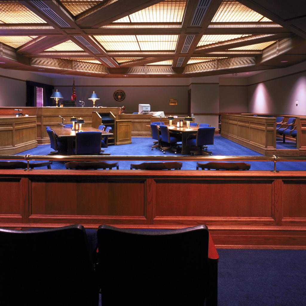 Court room interior with gridded ceiling lighting, wooden benches and tables, and blue carpeting