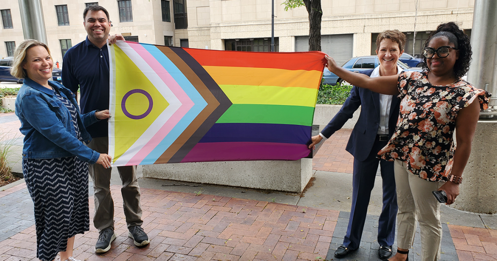Four people standing in front of a building holding the Progress Pride flag.