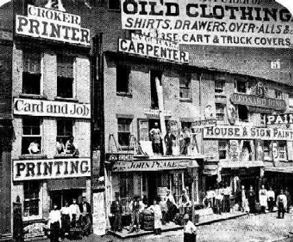 1865 photo of Lower Hudson Street, showing stacked storefronts for printing, clothing, paint, and a carpenter