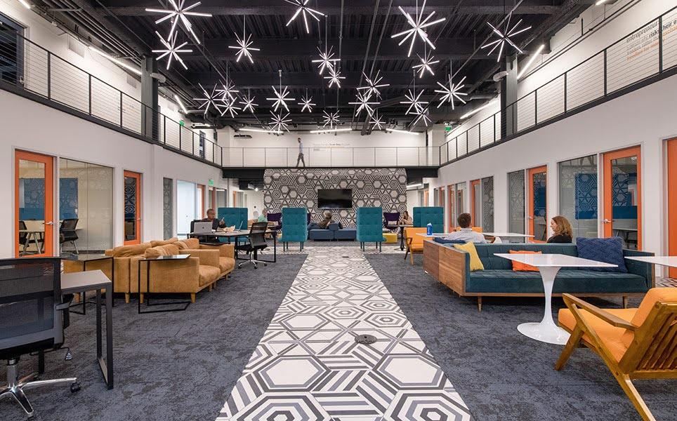 A large office space with starburst light fixtures and several people sitting on couches and armchairs, with a hexagonal patterned aisle.