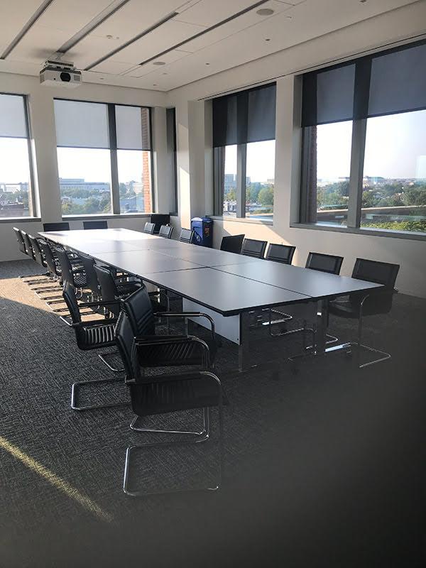 A long conference table with twenty office chairs in a room with a projector attached to the ceiling and several windows with shades.