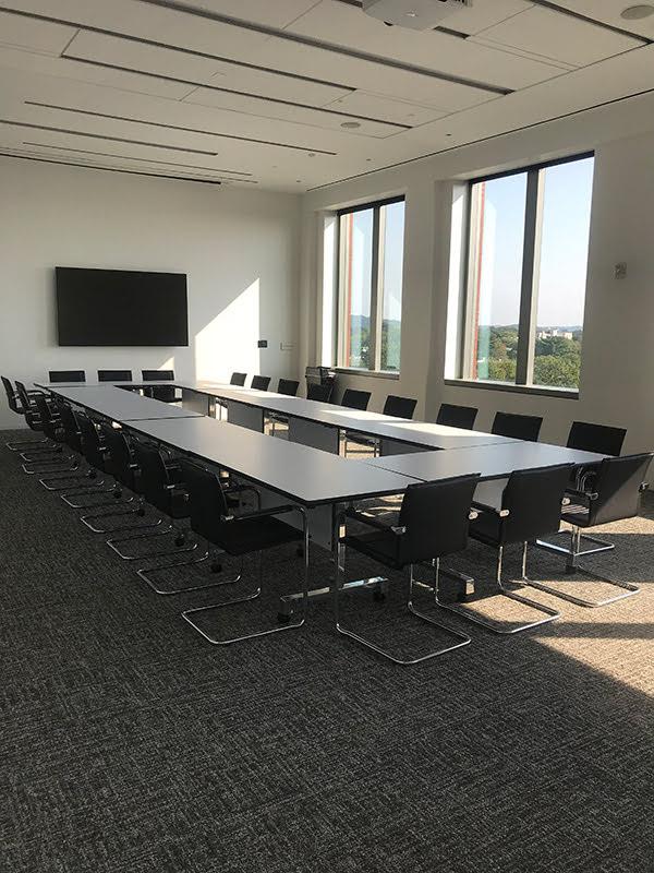 A carpeted conference room with conference tables in an elongated rectangle with multiple office chairs around and a flatscreen at far end.