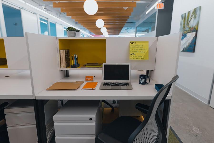 A white cube workstation with a shelf with notebooks, pens, and books, and a desk with a laptop, notebook, file folders, and a mug.