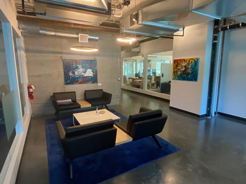 Concrete floors and walls with artwork on two walls and bench armchairs with tables and a blue rug underneath.
