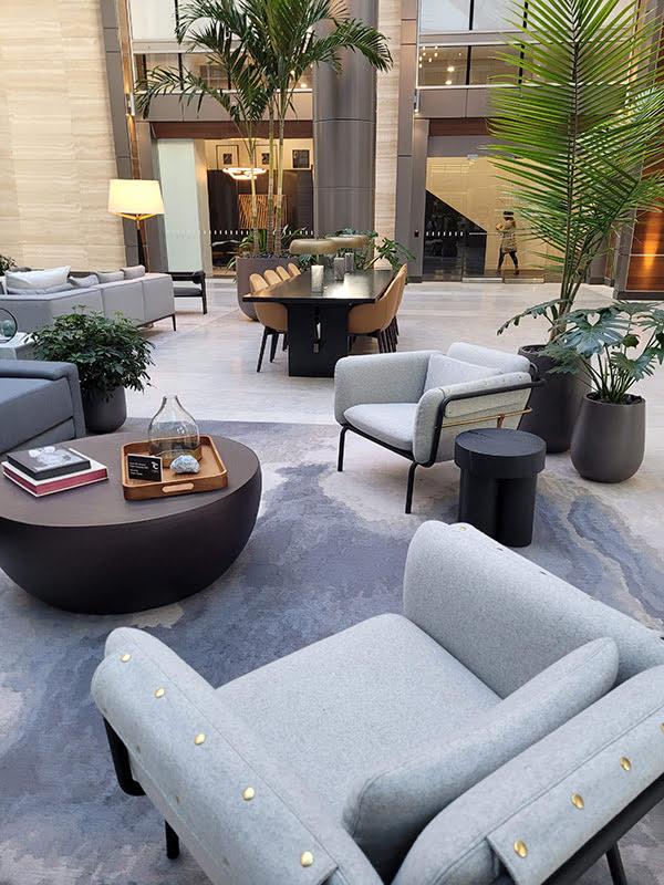 An open lobby area with several pieces of furniture, including two light gray, upholstered lounge chairs with round end tables and table.