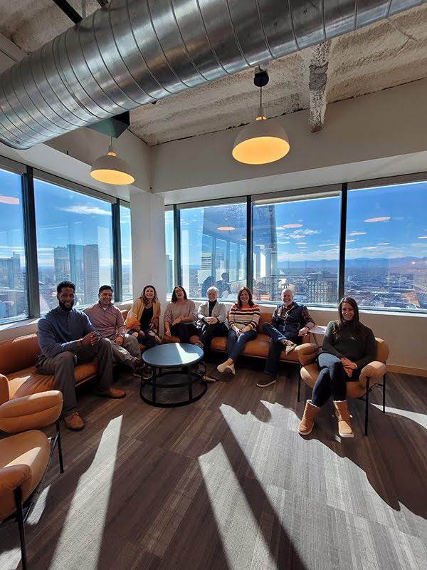 Several people sit on sofas and in armchairs in the corner of a carpeted office space with large windows behind them with buildings.