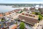 Aerial image of a four-story red brick building with a large one-story annex overlooking a parking lot, a row of brick buildings, streets, a harbor, and a bridge.