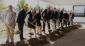 A row of 12 dignitaries hold shovels with dirt under a ceremonial tent outdoors on a job site.